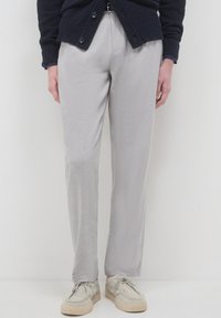 Persona che indossa pantaloni dritti grigio chiaro, scarpe beige con lacci e un cardigan blu scuro, in piedi davanti a uno sfondo bianco.