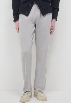 Persona che indossa pantaloni dritti grigio chiaro, scarpe beige con lacci e un cardigan blu scuro, in piedi davanti a uno sfondo bianco.