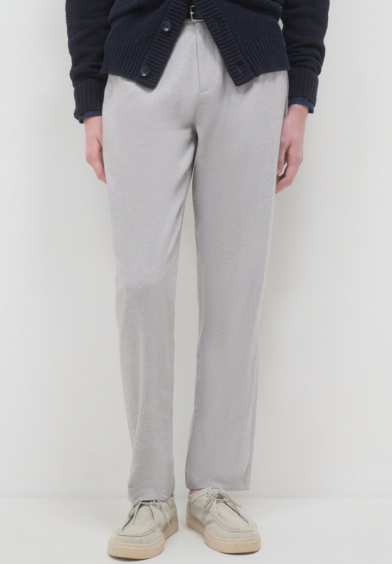 Persona che indossa pantaloni dritti grigio chiaro, scarpe beige con lacci e un cardigan blu scuro, in piedi davanti a uno sfondo bianco.