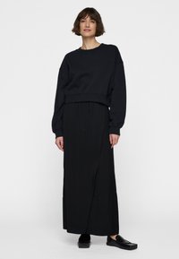 Sudadera negra recortada con mangas anchas combinada con una falda larga negra con cordón. Llevada con zapatos planos negros. Textura suave y mate.