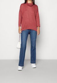 Bordeauxfarbene Cowl-Neck-Bluse mit langen Ärmeln, kombiniert mit blauen Schlagjeans. Accessoires: eine kleine weiße Schultertasche und weiße Sneakers.