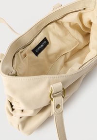 Sac à main beige avec un extérieur en similicuir lisse, une doublure intérieure en tissu, une fermeture zippée et une seule bandoulière avec des ferrures dorées.