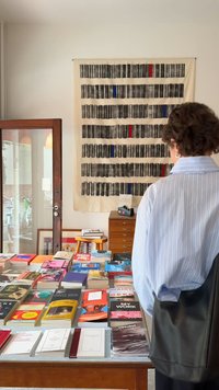 Galda attēlojums ar dažādām grāmatām ar krāsainām vākiem. Fons veido teksturēts sienas dekors ar melnām un sarkanām svītrām. Ir klāt persona.