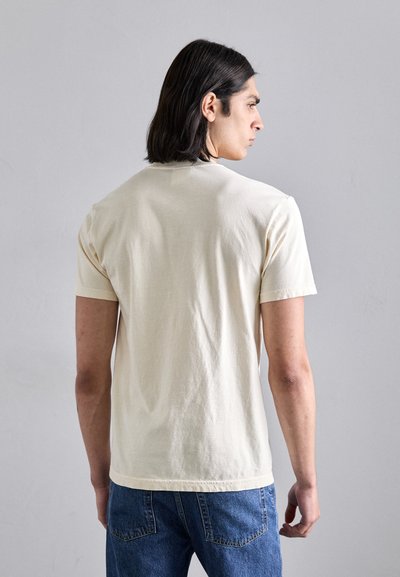 Light beige short-sleeve t-shirt with a crew neck, made from soft cotton. Features a plain design and a relaxed fit. Paired with blue jeans.