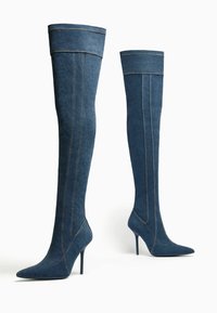 Džínové kozačky nad kolena s špičatou špičkou a jehlovým podpatkem. Mají kontrastní švy a těsný střih kolem lýtka. Barva je tmavě modrá.