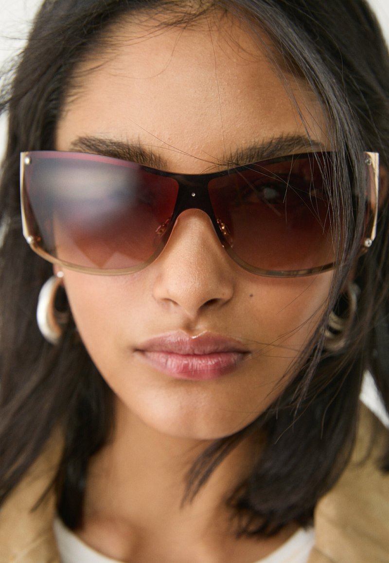 Sunglass med overdimensionerede mørkerøde linser og en transparent brun ramme, der har guldtonede accenter ved stængerne.