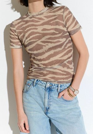 Personne portant un haut ajusté à manches courtes imprimé tigre marron et beige, avec un jean taille haute bleu clair et des bracelets dorés au poignet gauche.