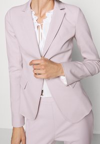 Vaaleanpunainen bleiseri, jossa on muotoiltu leikkaus, viisteet ja kaksi etutaskua. Päällä valkoinen paita, jossa on röyhelökaulus.