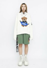 Sudadera blanca con un gráfico de un oso, pantalones cortos verdes, botas blancas y riñonera azul. Auriculares grises alrededor del cuello.