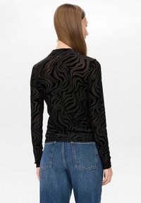 Frau mit glattem braunem Haar, die ein eng anliegendes, langärmeliges schwarzes Oberteil mit einem welligen Zebramuster und blauen Jeans trägt, mit dem Rücken zur Kamera.