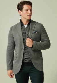 Grau blazer über einem gemusterten grauen Pullover mit Reißverschlusskragen, kombiniert mit dunkelgrünen Hosen; das Outfit wird durch ein Taschensquare mit blauem Design vervollständigt.