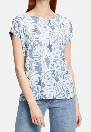 Femme portant un haut à manches courtes bleu et blanc à motif floral avec un col rond, et un jean bleu clair, se tenant avec une posture détendue.