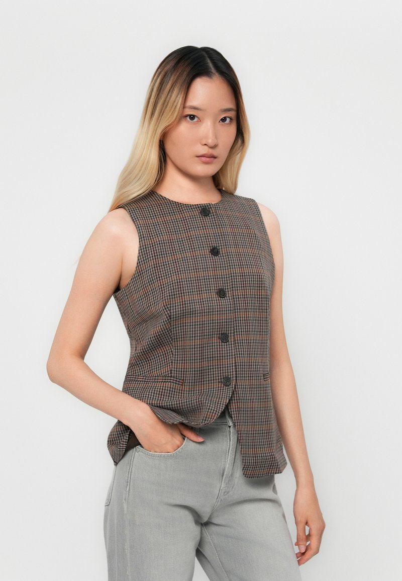 Frau mit langen Ombre-Haaren trägt eine ärmellose braun karierte Weste und hellgraue Jeans und posiert mit einer Hand in der Tasche vor einer weißen Wand.