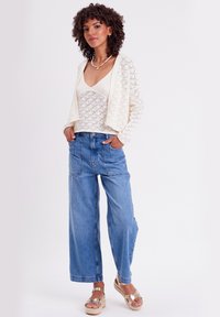 Cárdigan de ganchillo en color blanco roto, superpuesto a una blusa de color claro con textura de canalé. Combinado con pantalones de mezclilla anchos en azul y sandalias de cuña.