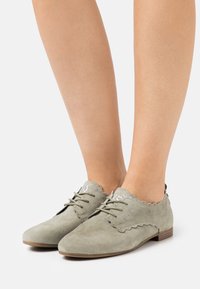 Zapatos oxford de ante verde con bordes ondulados, puntera redondeada y suela de madera plana. Los cordones en la parte frontal añaden un detalle funcional.