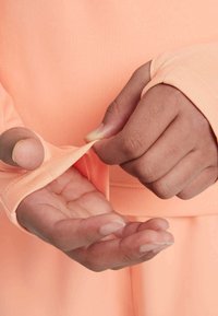 Długi rękaw w jasnoróżowym materiale, z otworami na kciuki w rękawach. Zbliżenie pokazuje dłonie trzymające tkaninę przy nadgarstku.