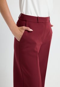 Pantalon bourgogne au tissu lisse, avec des poches latérales, des passants de ceinture et une coupe ajustée. La moitié d'un haut blanc est visible.