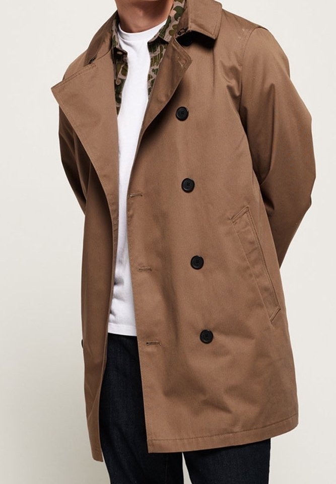 Homme portant un trench-coat beige à double boutonnage sur une chemise blanche avec col à motifs, mains derrière le dos, torse visible du menton aux cuisses.