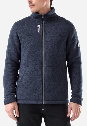 Man wearing zipped, dark blue fleece jacket with high collar and brand logo on chest and sleeve, standing against a plain white background.