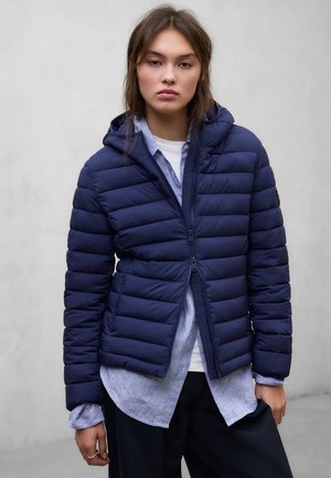 Ung kvinde iført navyblå polstret hættejakke over lyseblå skjorte og hvid top, stående mod en ensfarvet grå baggrund.