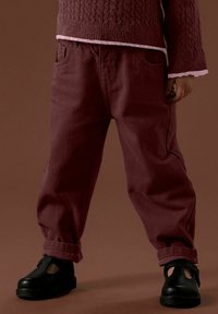 Bambino con pantaloni larghi color mogano arrotolati alle caviglie, maglietta a maniche lunghe di tessuto mogano con bordi rosa e scarpe nere con cinturini in piedi su un pavimento marrone.