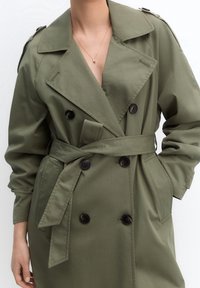 Oliwiona zielona trench coat wykonana z gładkiego materiału, z podwójnym zapięciem, dużymi czarnymi guzikami oraz zaciągniętym paskiem w talii.