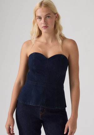 Blonde Frau trägt ein trägerloses dunkelblaues Denim-Top und passende dunkelblaue Denim-Jeans vor einem schlichten hellen Hintergrund.