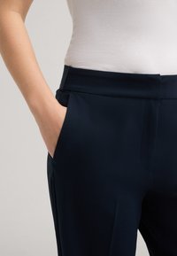 Marineblaue Stretchhosen aus glattem Stoff mit flachem Bund und Seitentaschen, teilweise mit einem weißen Oberteil dargestellt.