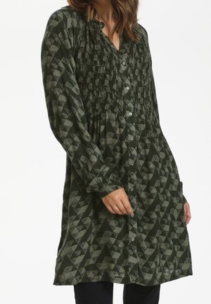 Femme portant une robe-tunique boutonnée à manches longues avec un motif géométrique texturé vert foncé et gris.