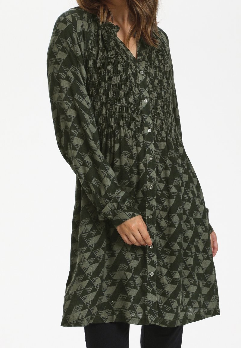 Femme portant une robe-tunique boutonnée à manches longues avec un motif géométrique texturé vert foncé et gris.