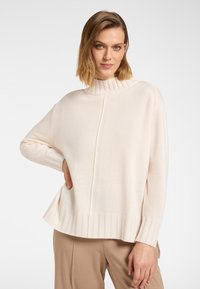 Frau mit hellbraunen Haaren, die einen cremefarbenen Strickpullover mit hohem Kragen und beige Hosen trägt und mit einer Hand in der Hüfte posiert.