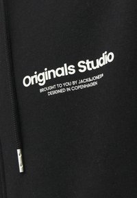 Zwarte hoodie van zacht materiaal met een ritssluiting aan de voorkant, een trekkoord en witte tekst: "Originals Studio" met merkdetails eronder.