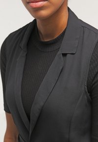 Černá na míru šitá vesta s výrazným střihem a klopami přes černý žebrovaný rolák. Hladká textura, přiléhavý vzhled a čisté linie.