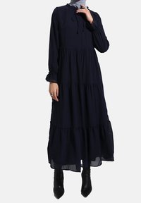 Lang marineblå kjole lavet af letvægtsstof, med lange ærmer, et lagdelt design og en indsnøring med rynker i halsudskæringen.