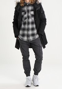 Svart parkas med pälsfodrad huva över en grå rutig skjorta, kombinerad med grå cargobyxor och vita sneakers. Synliga fickor på byxorna.