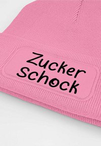 Pink knit beanie featuring a rectangular patch with the text "Zucker Schock" in black, highlighting textured fabric and ribbed pattern.