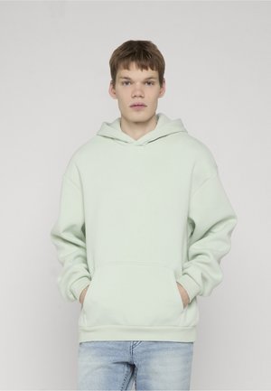 Fiatal férfi világosbarna hajjal, világoszöld kapucnis pulóvert és világoskék farmert viselve, kezét a pulóver zsebében tartva, egyszerű háttér előtt áll.