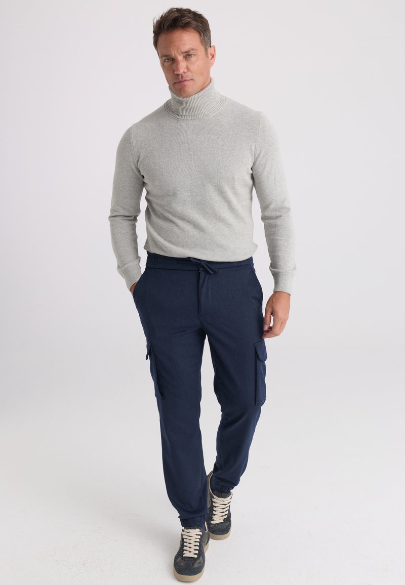 Homme portant un pull à col roulé gris clair, un pantalon cargo bleu marine et des baskets noires marchant devant un fond blanc uni.