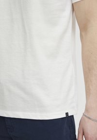 Hvit bomulls t-skjorte med korte ermer, med et lite svart merke på nederste kant. Stoffet har en glatt tekstur og en løs passform.