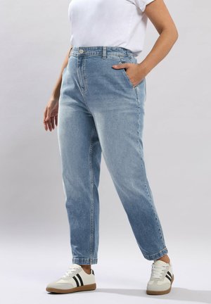 Person iført lyseblå højtaljede jeans, hvid t-shirt og hvide sneakers med sorte striber, stående med den ene hånd i lommen.