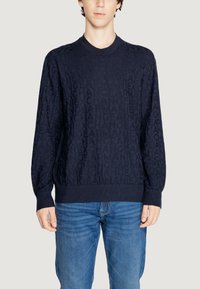 Maglione blu navy caratterizzato da un motivo testurizzato di lettere ripetute, scollo rotondo e polsini e vita a costine. Abbinato a jeans blu.