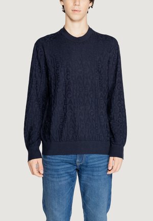 Maglione blu navy caratterizzato da un motivo testurizzato di lettere ripetute, scollo rotondo e polsini e vita a costine. Abbinato a jeans blu.