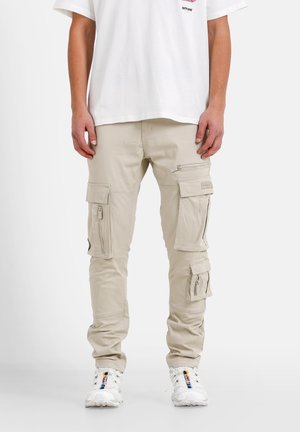 Beige cargobroek met meerdere zakken, ritsen en een aangesloten ontwerp, gecombineerd met een losvallend wit T-shirt en witte sneakers.