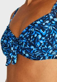 Blå blommig bikiniöverdel med rynkdetalj, justerbara remmar och ett dekorativt band framtill. Klara färger mot en mörk bakgrund.