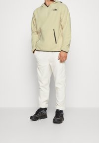 Person som bär en beige hoodie och vita sweatpants med svarta vandringsskor, står framför en enfärgad ljusgrå bakgrund.