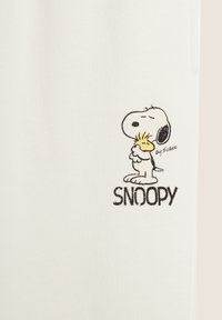 Tissu blanc ornée d'un design brodé noir de Snoopy tenant un objet jaune, avec "SNOOPY" en texte noir gras sous l'illustration.