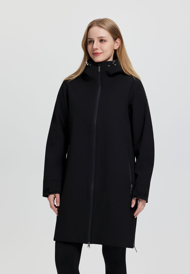 Kvinde iført en lang sort frakke med lynlås, høj krave og sidelommer med lynlås, stående mod en ensartet lys baggrund.