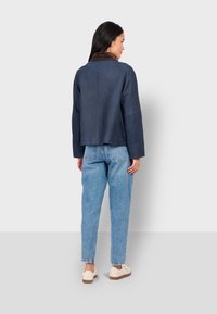 Námořnická modrá zamšová bunda s hnědým límcem, uvolněný střih, spadlé ramena a zkrácená délka, kterou doplňují světle modré džíny a krémové tenisky.