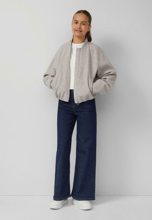 Jonge vrouw in een lichtgrijze zipjack, witte shirt, donkerblauwe wijde jeans en witte sneakers, staand tegen een grijze muur.