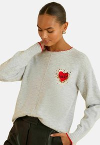 Grauer Pullover mit einem roten Herz-Patch und dem Text "With love", verziert mit goldenen Pailletten. Rote Ripp-Details am Kragen und an den Ärmeln.